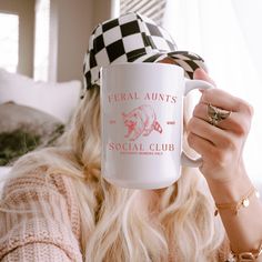 a woman with long blonde hair holding a coffee mug in front of her face and wearing a checkered hat