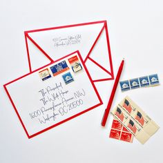 two envelopes with stamps and a red pen are on a white surface next to some postcards