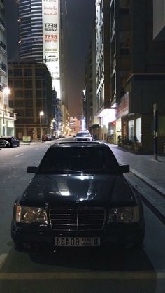 a black car parked on the side of a city street at night with tall buildings in the background