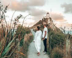 a man and woman walking down a path towards the ocean with a lighthouse in the background