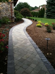 a brick walkway in front of a house with flowers and trees on the other side