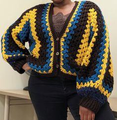 a woman standing in front of a white wall wearing a colorful crocheted jacket