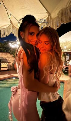 two beautiful young women hugging each other in front of a swimming pool at night time