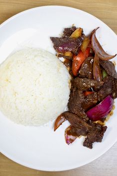 a white plate topped with meat and rice on top of a wooden table next to a fork