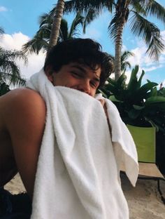 a man wrapped in a white towel sitting on the beach with palm trees behind him