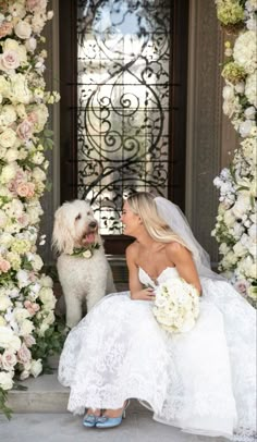 a woman in a wedding dress sitting next to a white dog on the steps with flowers all around her