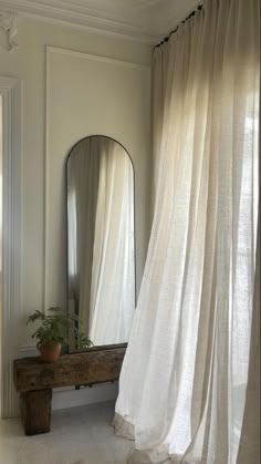 a mirror and bench in a room with white curtains on the windowsill, along with a potted plant