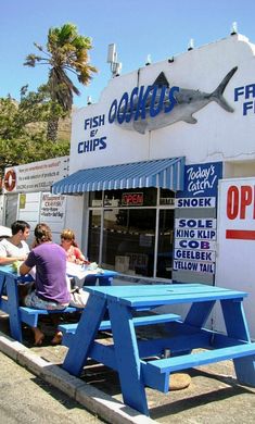 people sitting at blue picnic tables in front of a fish and chips shop with a shark on the wall