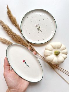 two white plates with designs on them are being held by someone's hand next to some dried grass