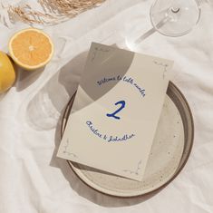 a table topped with oranges and paper next to a glass of wine on top of a white cloth covered table