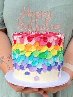 a person holding a birthday cake with rainbow icing on it and the words happy birthday written on top