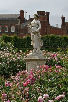 there is a statue in the middle of some flowers and bushes with a building in the background