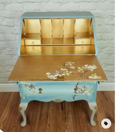 an antique desk painted in blue and gold with flowers on the top, sitting against a white brick wall