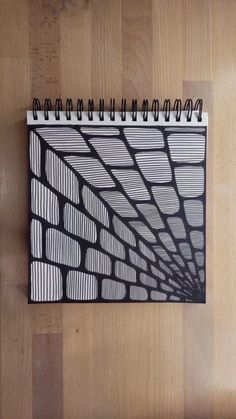 a spiral notebook with black and white lines on it sitting on a wooden floor next to a pen