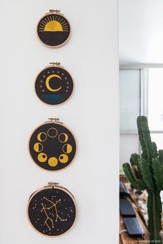 three hand embroidered wall hangings on a white wall next to a potted cactus