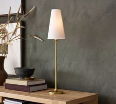 a lamp on a table next to a mirror and some books in a room with grey walls