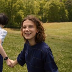 two young children holding hands in a grassy field with trees in the background and one child smiling at the camera