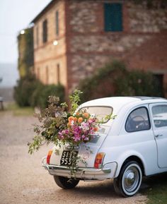 an old car with flowers in the back parked on a gravel road next to a brick building