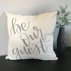 a white pillow with the words be our guest on it next to a black vase