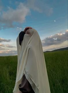 a woman standing in the grass covering her face with a white blanket over her head