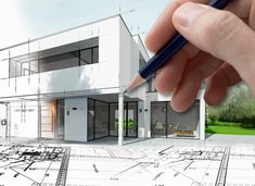 a hand holding a pencil drawing a house with blueprints on the ground and trees in the background