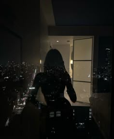 a woman standing in front of a window at night with the city lights behind her