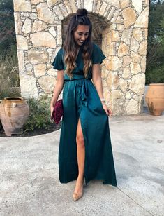 a woman standing in front of a stone wall wearing a green dress and holding a purse