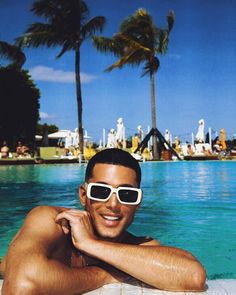 a man wearing sunglasses sitting on the edge of a swimming pool with palm trees in the background