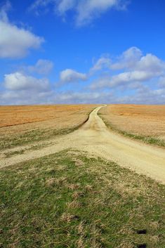 a dirt road in the middle of an open field with grass on both sides and blue skies above