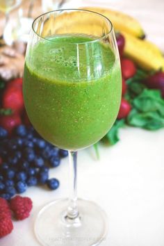 a green smoothie in a wine glass surrounded by berries and bananas