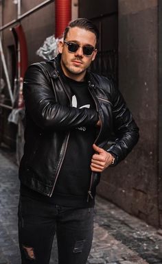 a man in black leather jacket and sunglasses standing on the street with his arms crossed