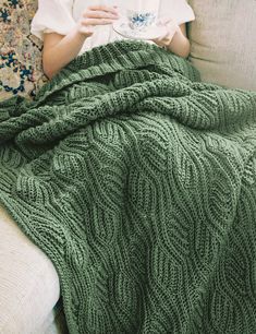 a woman sitting on a couch holding a cup in her hand while wrapped up in a blanket