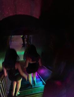 three women are sitting on an escalator in the dark, with neon lights