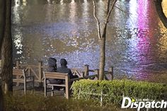 two people sitting on a bench looking at the water in front of some trees and bushes