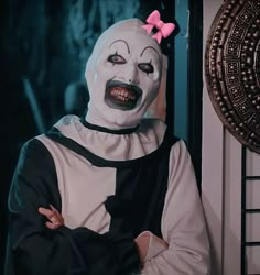 a person wearing a creepy mask and pink bow standing in front of a door with their arms crossed