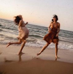 two women are running on the beach near the ocean at sunset or sunrise, one is wearing a brown dress