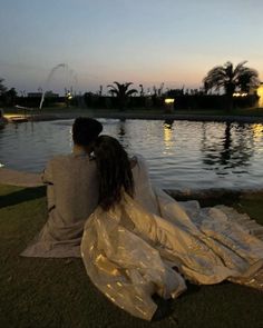 two people sitting on the grass next to a body of water at sunset or dawn