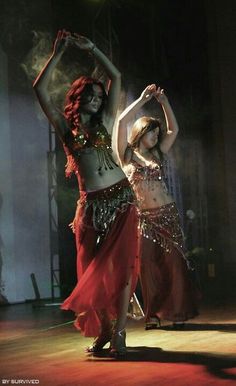 two women are dancing on stage with their arms in the air and one is wearing a belly dance costume