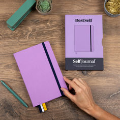 a person's hand is writing on a purple notebook next to some pens and pencils