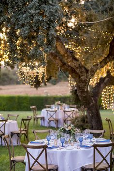 an outdoor table set up for dinner under the shade of a tree with string lights