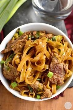 a white bowl filled with noodles and meat on top of a wooden table next to green onions