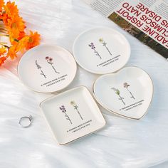 three white plates with flowers on them sitting next to an orange flower and a book