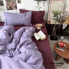 a bed with purple sheets and pillows next to a small table with an open book on it