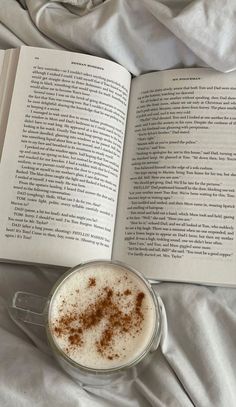 an open book sitting on top of a bed next to a cup of coffee with cinnamon sprinkles