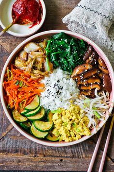 a bowl filled with rice, vegetables and meats next to chopsticks on a wooden table