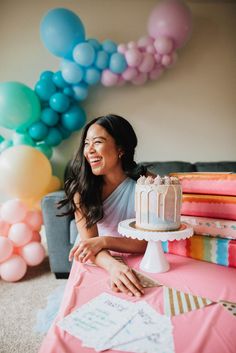 a woman sitting at a table with a cake in front of her and balloons behind her