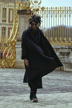 a man with dreadlocks walking in front of a golden gate wearing a black coat