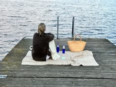 a woman sitting on top of a wooden dock next to a basket and bottles near the water