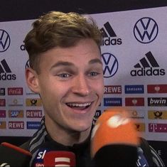 a man holding a microphone in front of a wall with soccer balls on it and talking to the media