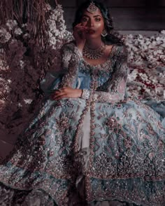 a woman in a blue and silver dress sitting on a bed of flowers with her hands to her face